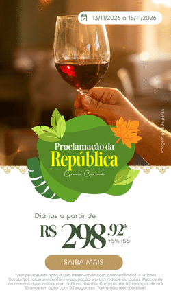 Close de uma mão segurando uma taça de vinho tinto em um ambiente elegante e iluminado do Grand Carimã Resort. Banner promocional da Proclamação da República com diárias a partir de R$ 298,92.